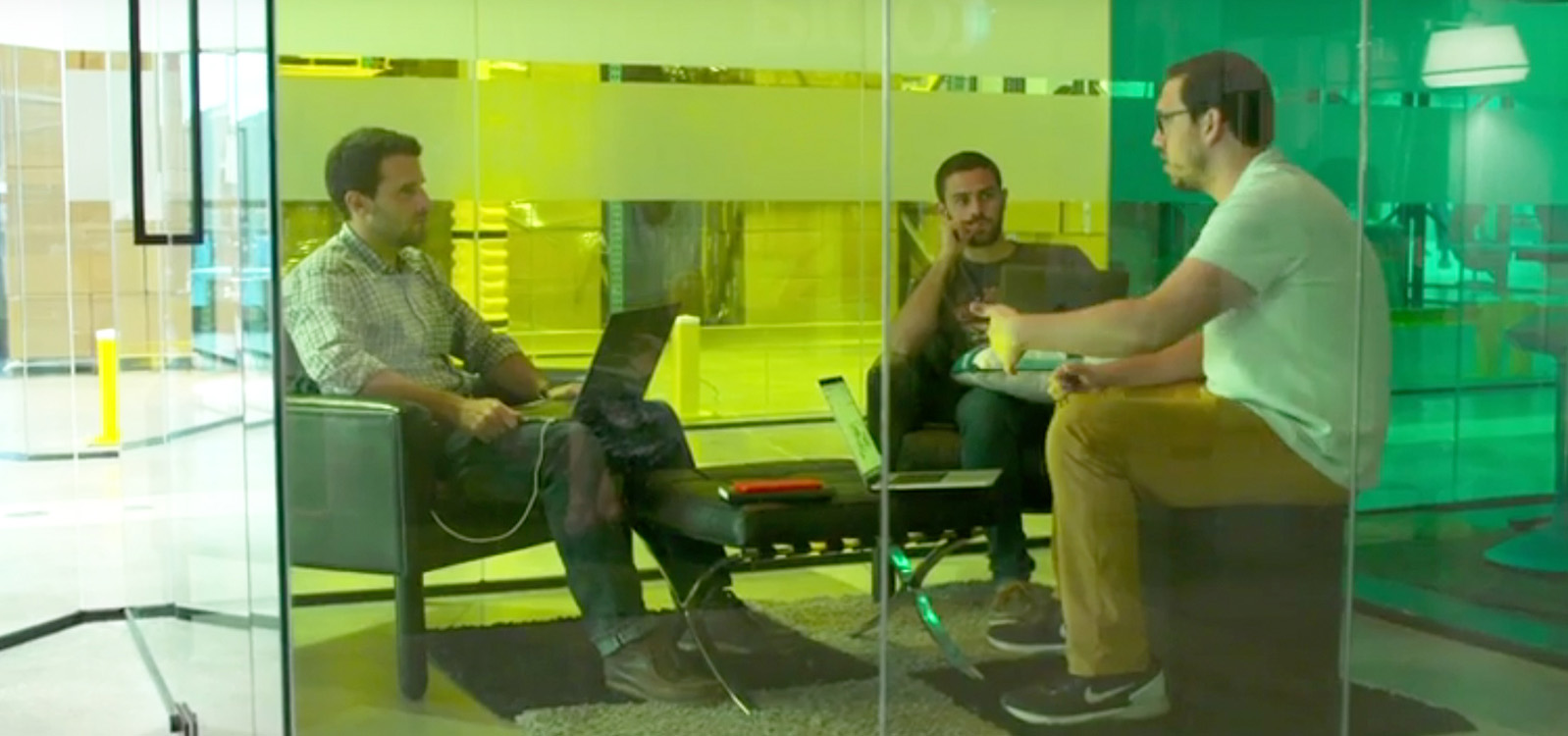 Três homens em uma reunião em uma sala de reuniões pequena com paredes de vidro